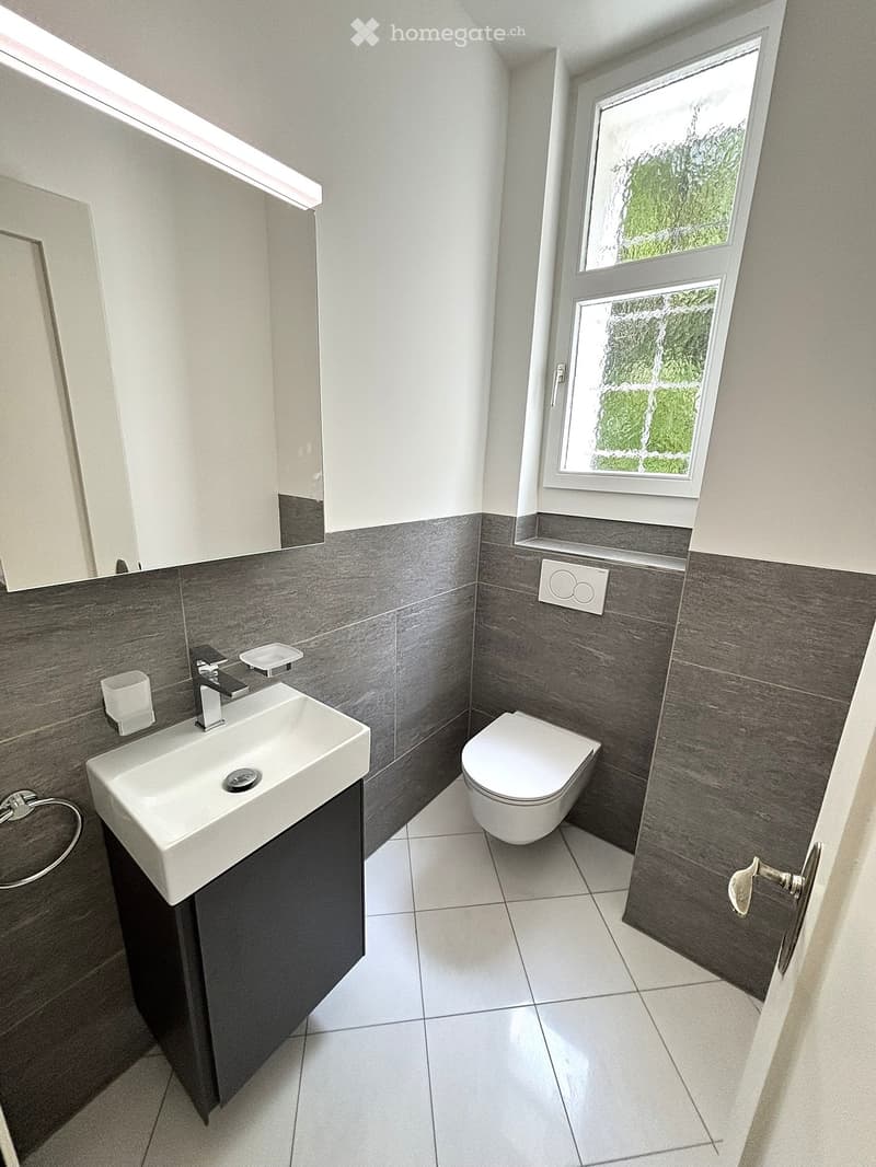 Modern bathroom with a white sink, toilet, and dark grey tiles on the walls. Features a window with frosted glass and a sleek faucet.