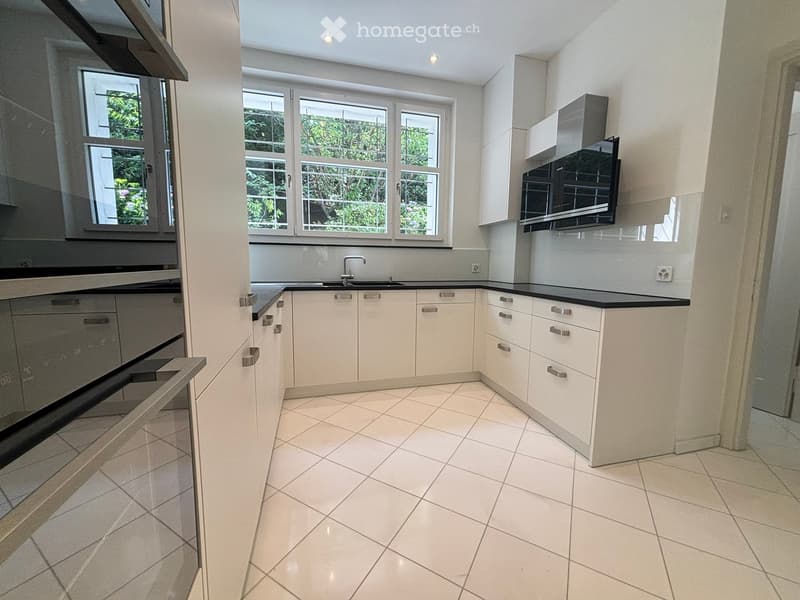 Modern kitchen with white cabinets, black countertops, and tiled flooring. Features a sink with a modern faucet and large windows offering a view of greenery.