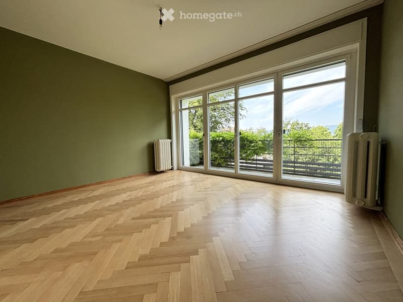 Empty room with wooden herringbone flooring, large sliding glass doors leading to a balcony, and radiators on both sides.