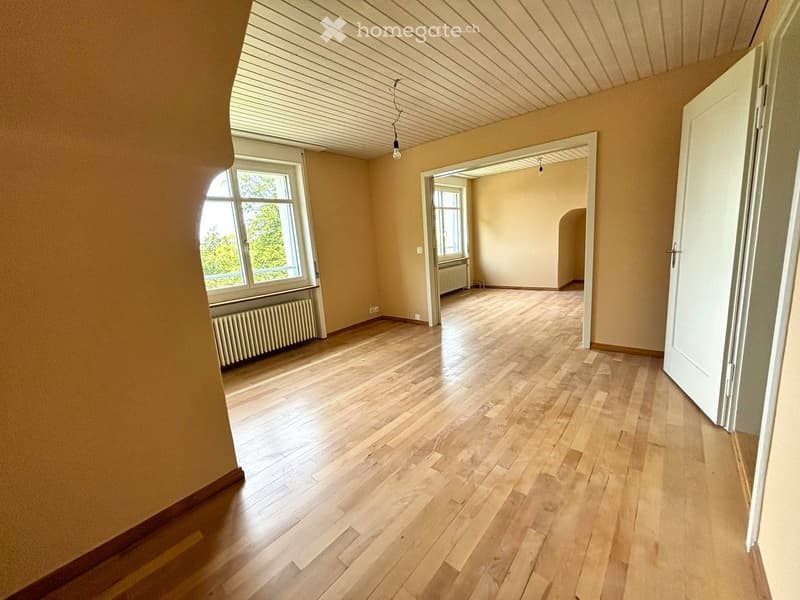 Empty room with wooden flooring, light-colored walls, and large windows allowing natural light. There are no visible furnishings or decorations.
