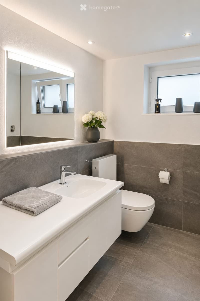 Modern bathroom with a white sink, chrome faucet, toilet, large mirror, and a vase with white flowers on the counter. Grey tiles on lower walls and dark brown tiles on the floor.