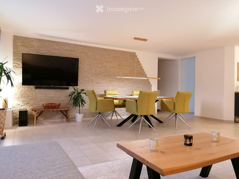 Modern living room with a large flat-screen TV mounted on a stone wall, a wooden coffee table with decorative candles, a dining area with yellow chairs and a table, a small bench with a basket, and a potted plant.