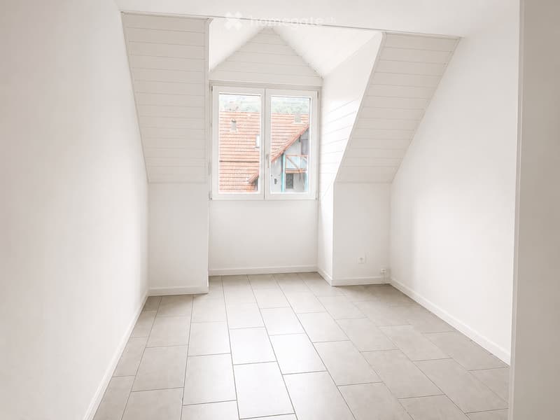 Empty room with white walls, tiled floor, and a large window offering a view of a red-roofed building.