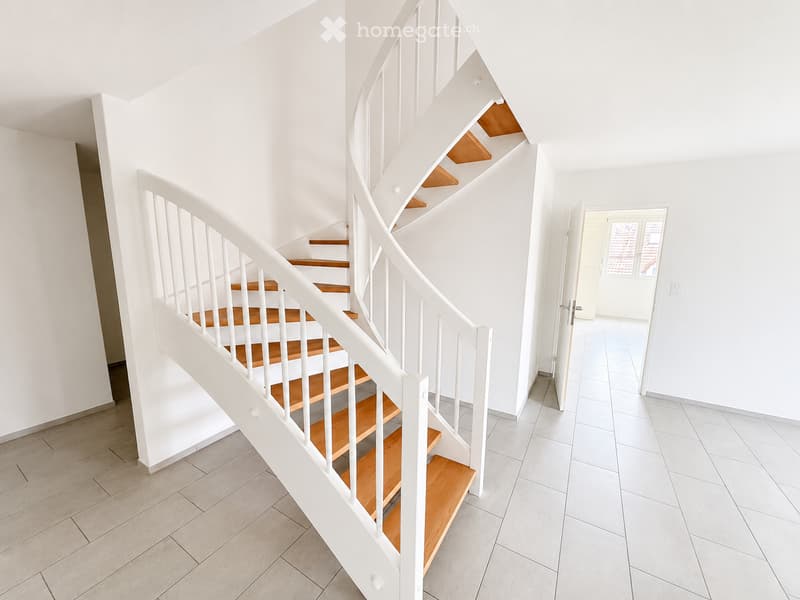 Spiral staircase with wooden steps and white railings, leading to an open doorway with white tiled flooring and walls.