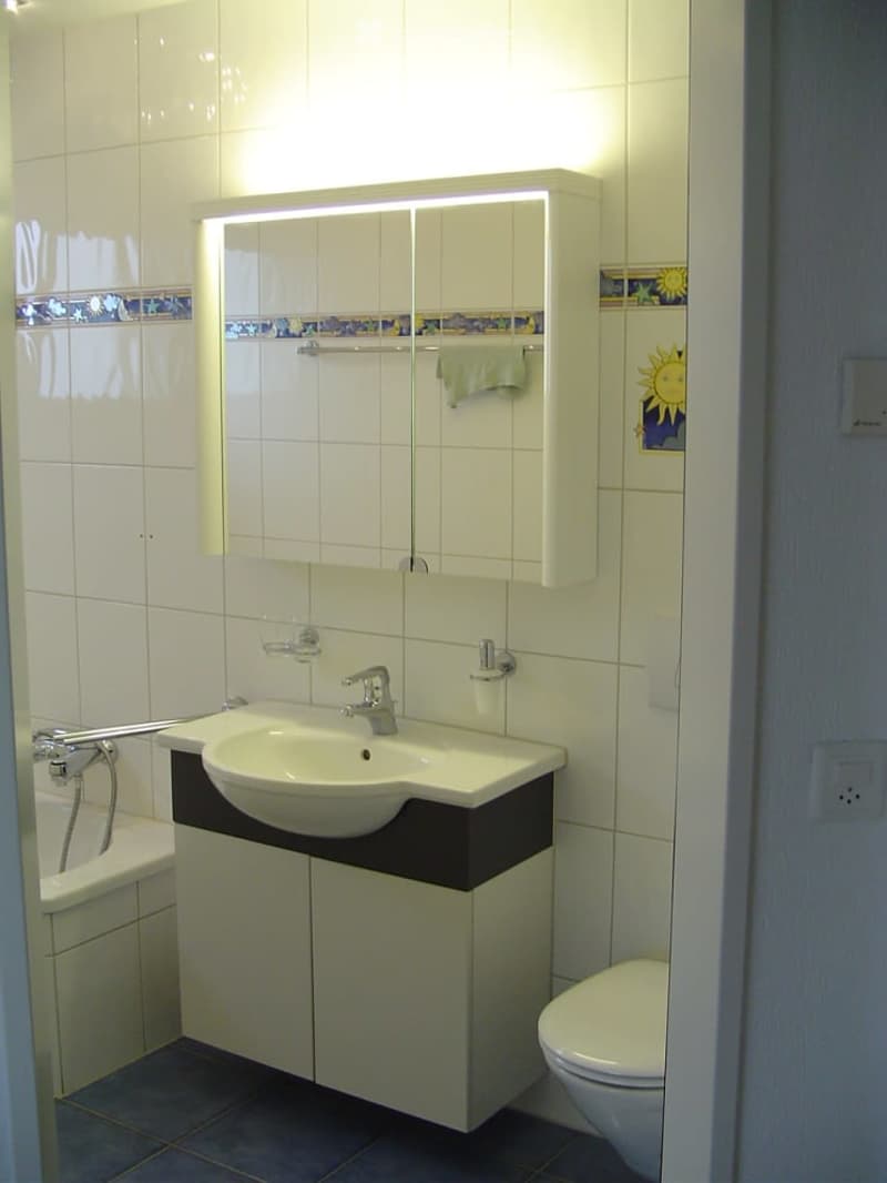 Modern bathroom with white tiles, a sink with a mirror cabinet, a toilet, and a bathtub. The mirror has built-in lighting, and the walls feature decorative tiles near the top.
