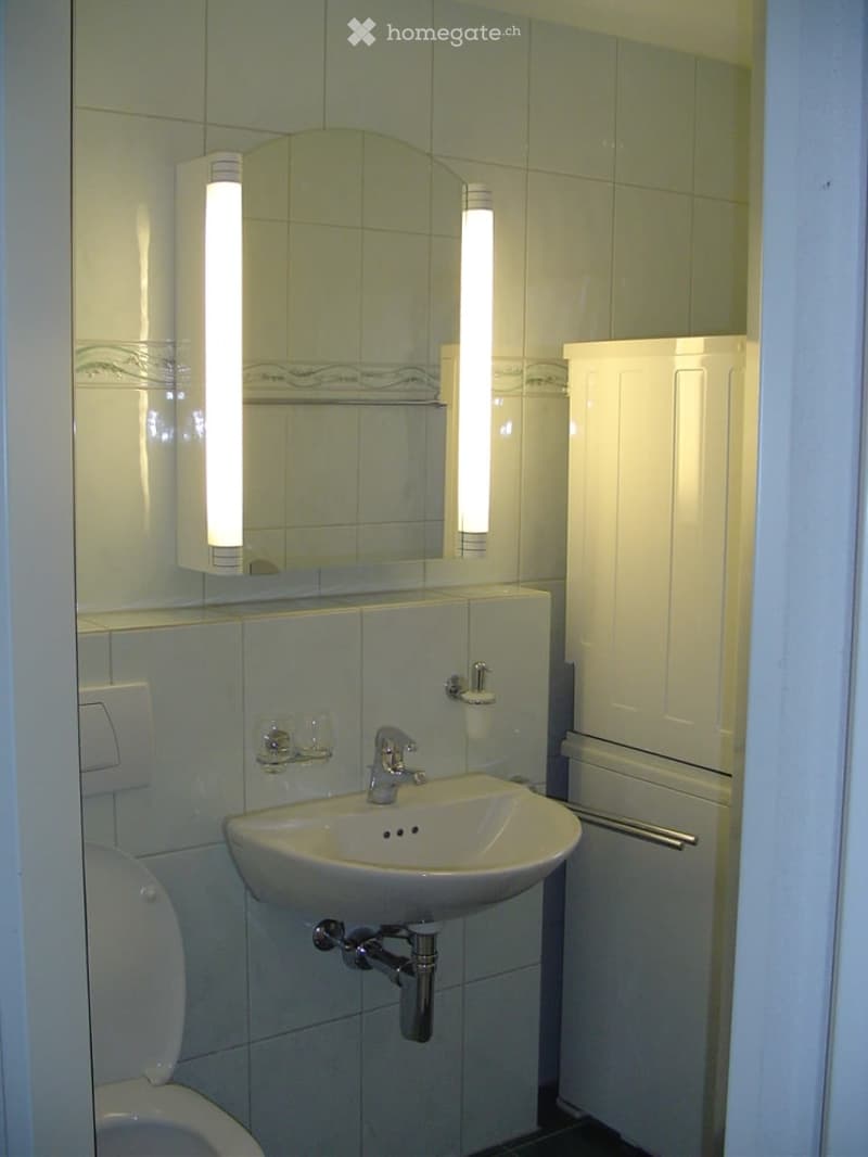 Modern bathroom with white tiles, a sink with a chrome faucet, a toilet, a mirror with built-in lighting, and a white cabinet.