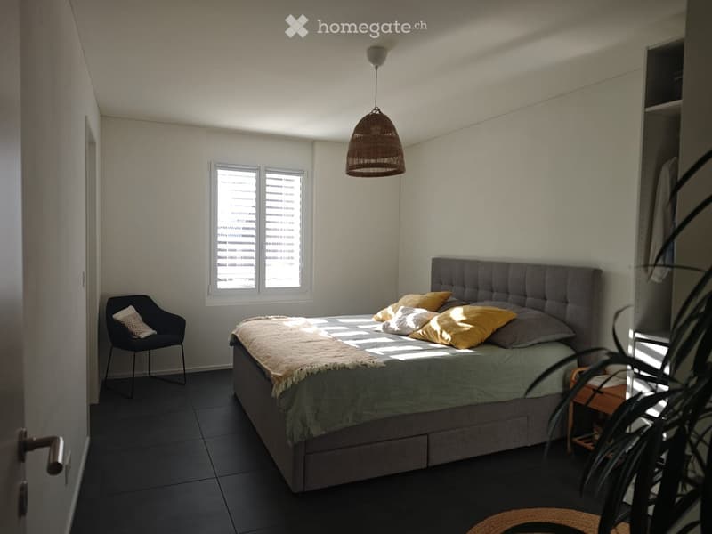 Modern bedroom with a grey upholstered bed, yellow and white pillows, a dark grey chair, and a woven pendant light hanging from the ceiling.
