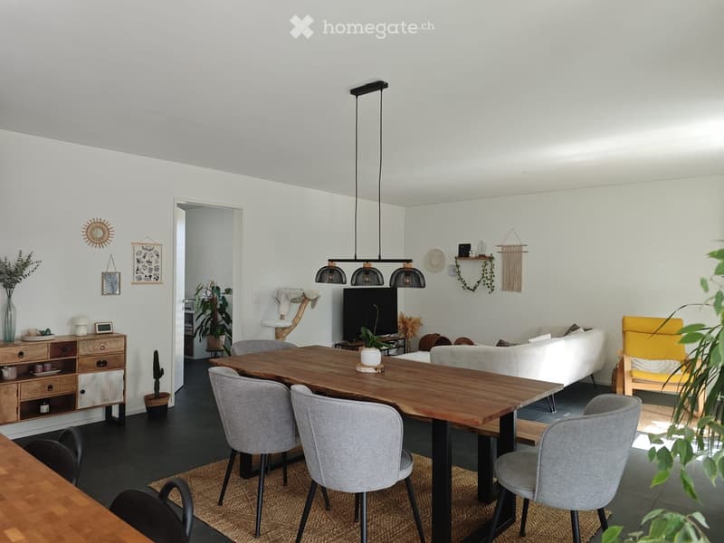 Bright living room with a wooden dining table, grey chairs, a white sofa, a television, and decorative plants.
