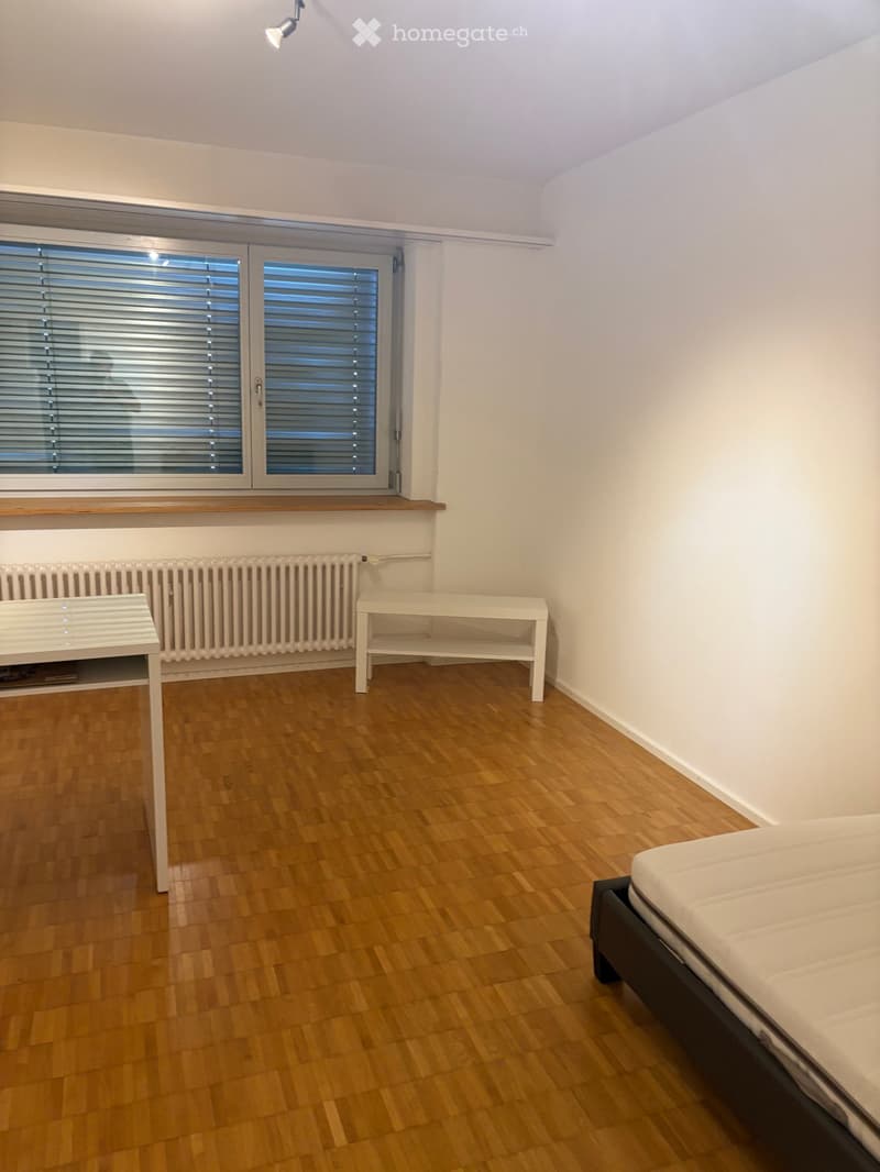Minimalist bedroom with wooden flooring, white walls, a single bed, small table, radiator, and large window with blinds.