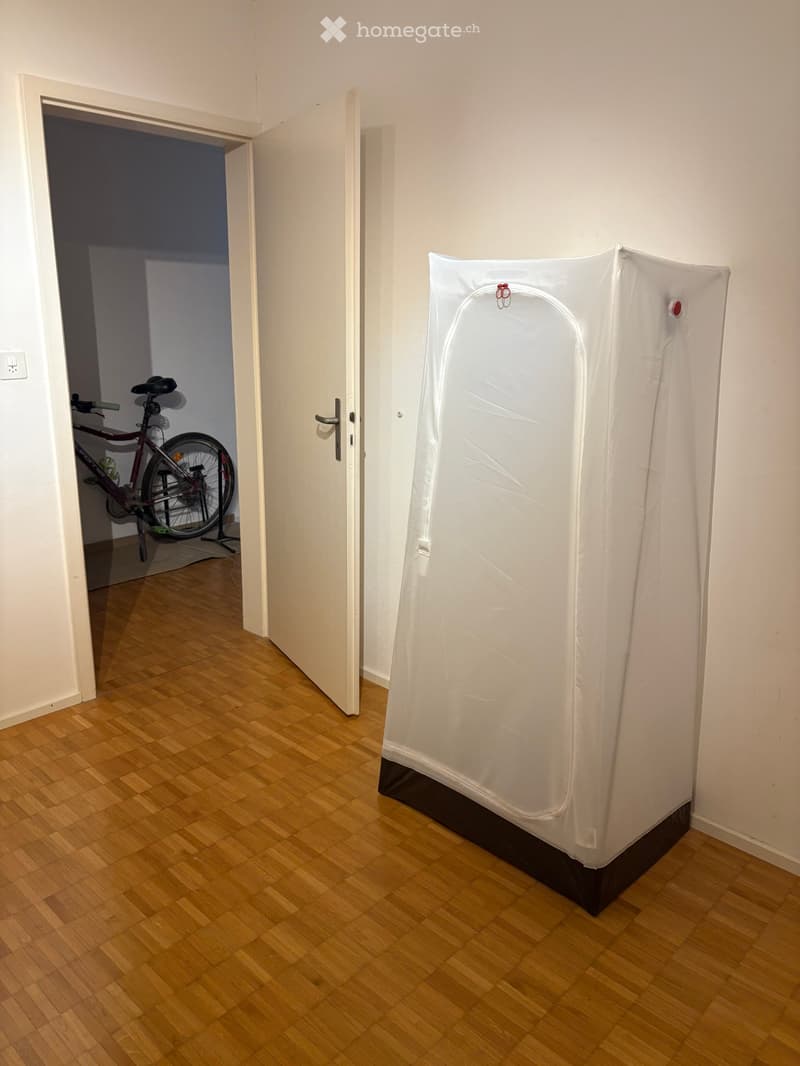 Empty room with wooden flooring, white walls, and a partially open door leading to another room with a bicycle inside.