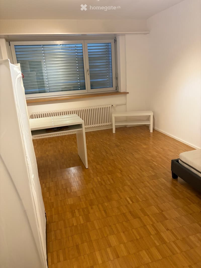 A small, unfurnished room with wooden flooring, a window with blinds, a radiator, and a bench. The room is partially visible with a bed in the corner.