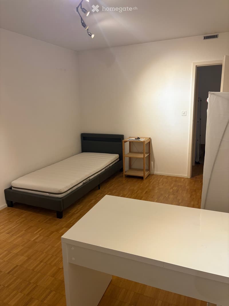 A small, furnished bedroom with a wooden floor, a bed with a black headboard, a small wooden shelf, and a white table.