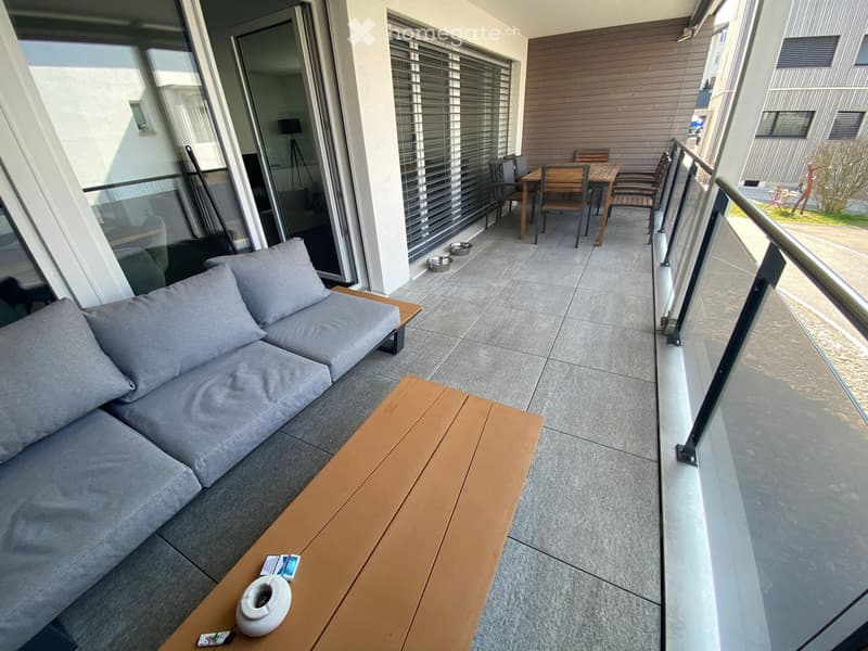 Balcony with grey sofa, wooden coffee table, dining table with chairs, and pet bowls.