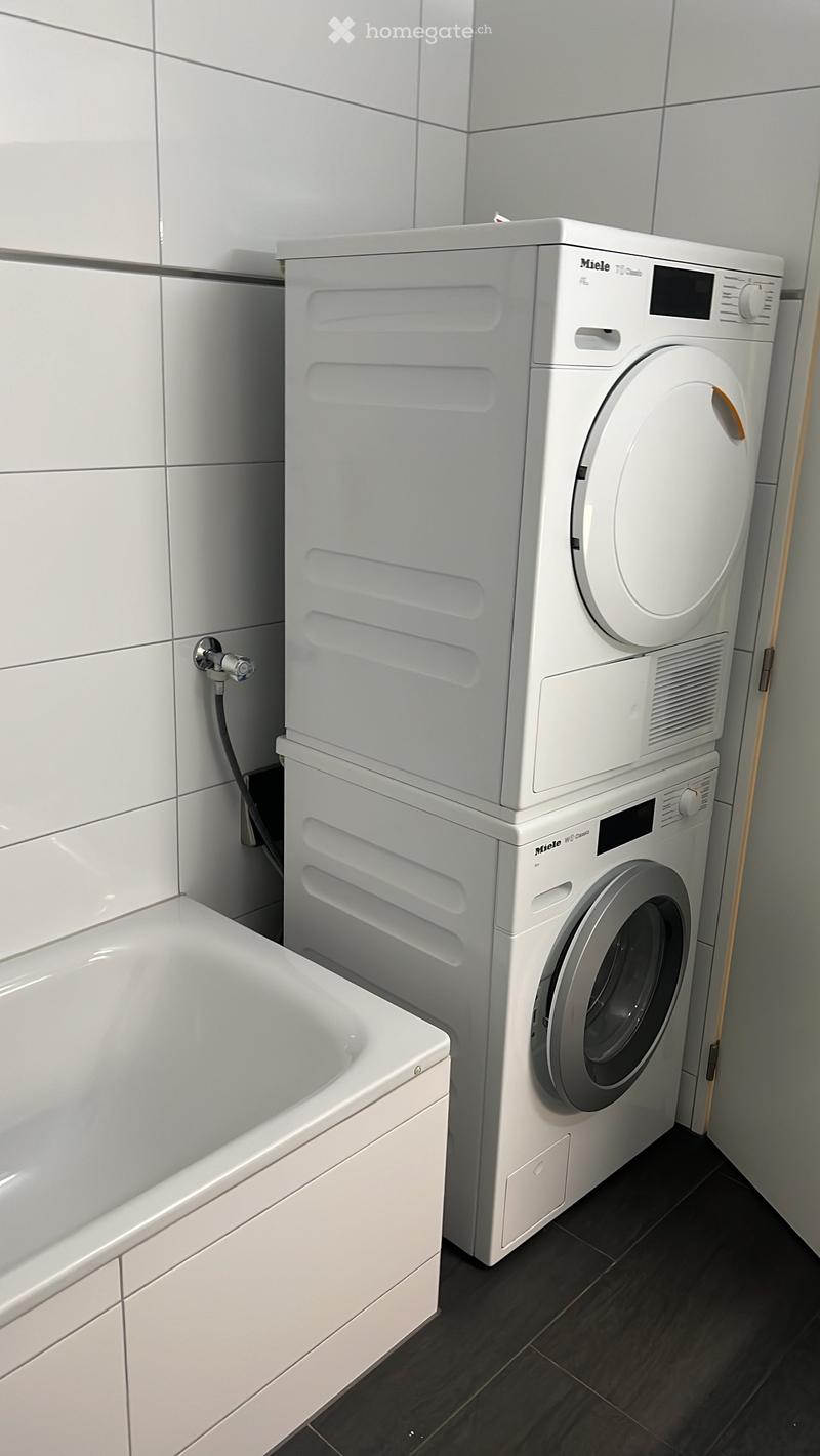 Bathroom with a bathtub, a washing machine, and a dryer. The washing machine and dryer are stacked vertically, and the bathtub is positioned to the left.