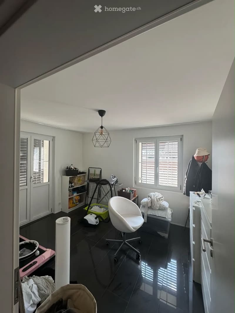 A modern, well-lit room with a white chair, a shelf, a small table, a mirror, a hanging lamp, a cabinet, and some folded clothes on a surface. The room has large windows with blinds and a dark tiled floor.