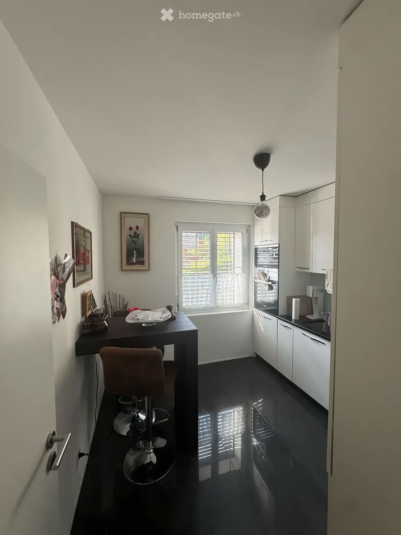 A modern kitchen with white cabinets, black countertops, and a small dining area with a table and stools. The room features a large window with blinds, a hanging light fixture, and framed pictures on the wall.
