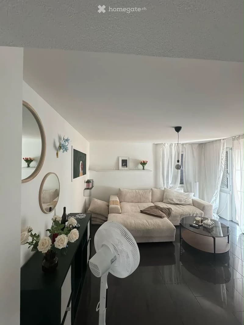 Modern living room with a beige sofa, glass coffee table, black side table with flowers, and a white fan. The room features large windows with white curtains, wall-mounted mirrors, and decorative wall art.