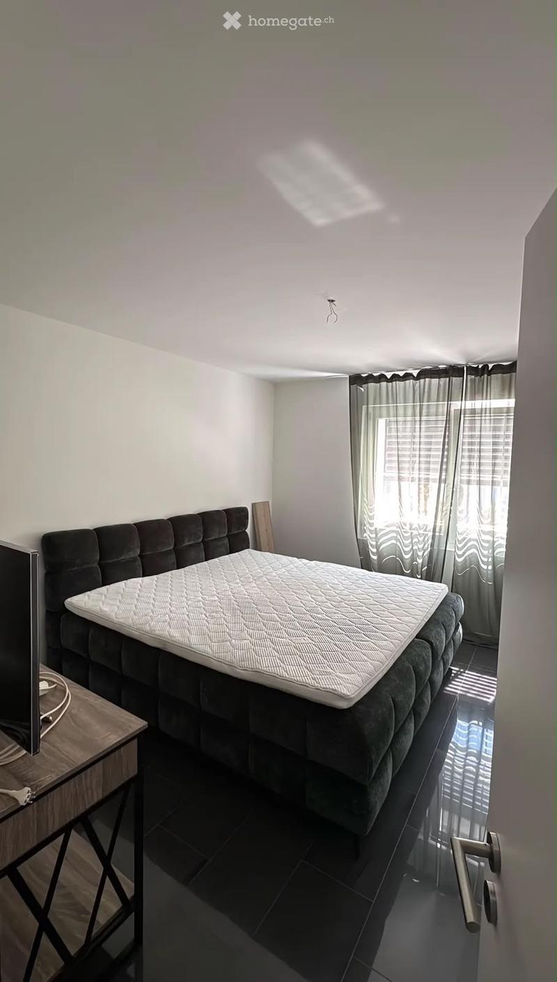 Modern bedroom with a dark upholstered bed, a white mattress, a wooden nightstand, and sheer curtains on the window.