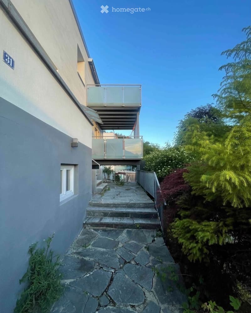 A narrow stone-paved pathway leads up to a modern building with a glass balcony and stairs.