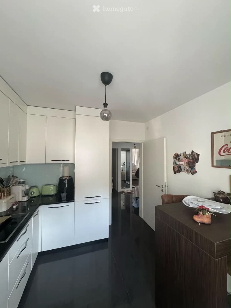 Modern kitchen with white cabinets, black countertops, and various appliances. A small dining area with a table and chairs is visible through an open doorway.