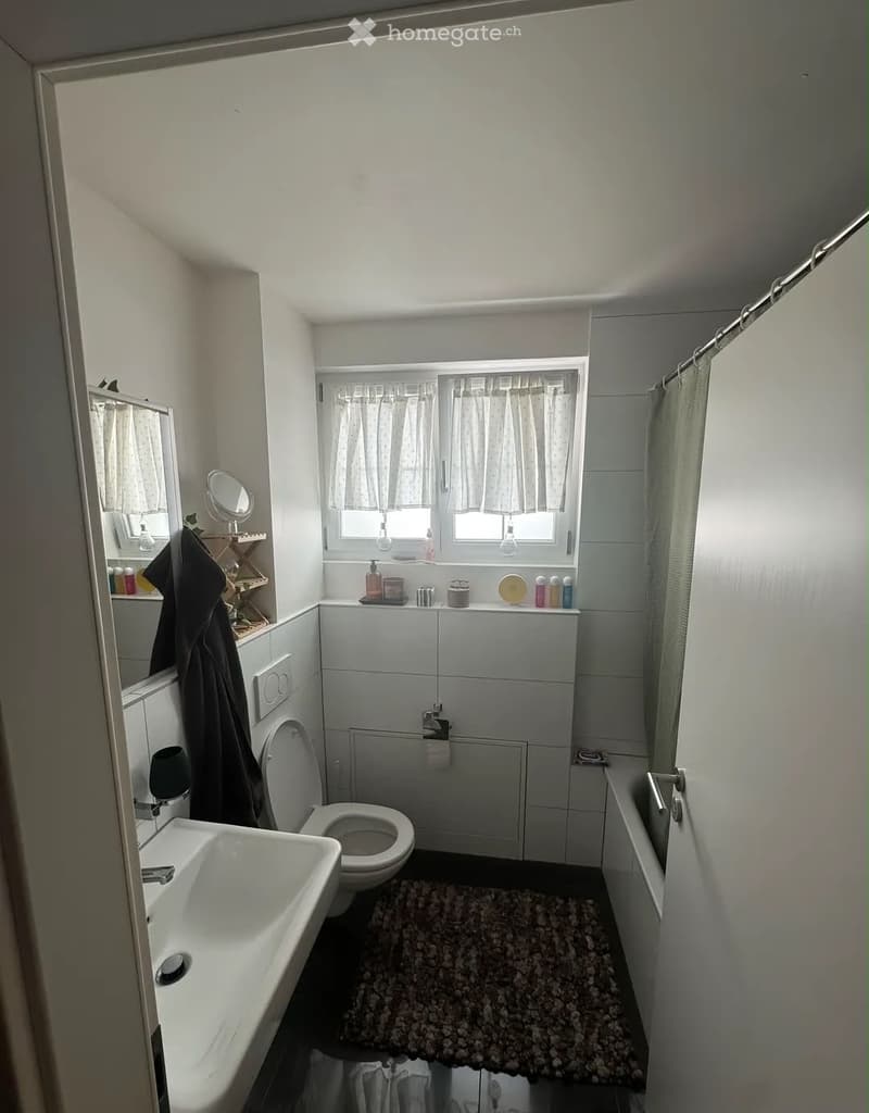 A compact bathroom with a white sink, toilet, bathtub, and a small window with curtains. A towel hangs on the wall, and a rug is placed in front of the bathtub.