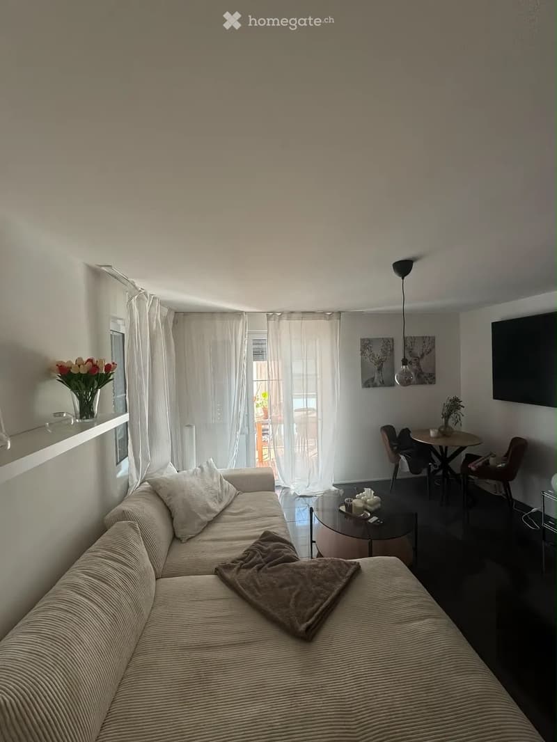 Modern living room with a beige sofa, coffee table, dining area with chairs and table, large windows with white curtains, and a television mounted on the wall.