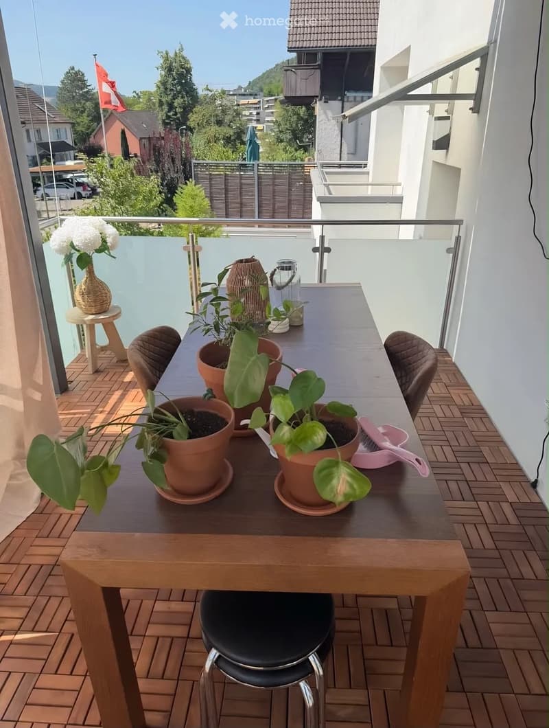 Balcony with wooden floor, glass railing, table with chairs, potted plants, vase, and decorative items.