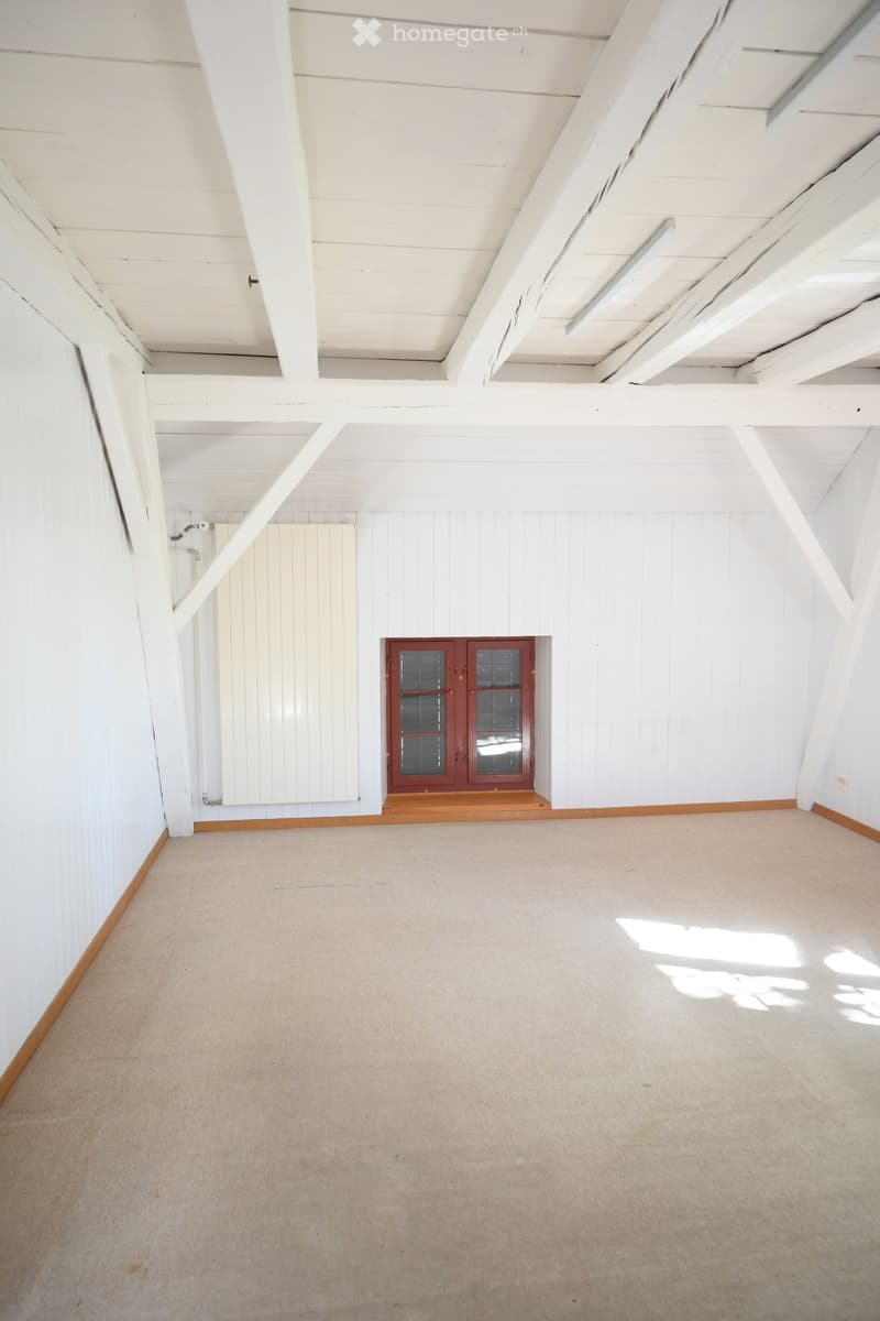 Empty room with white wooden walls, exposed wooden beams on the ceiling, and a single window with red framing. The floor is carpeted in a light beige color.