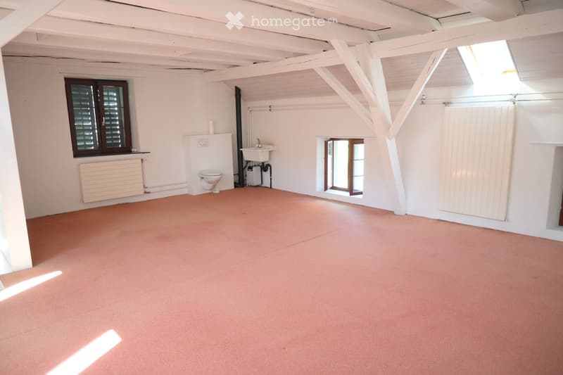 Empty room with white walls, exposed wooden beams, and a slanted ceiling. Features include a toilet, sink, radiator, and windows with shutters.