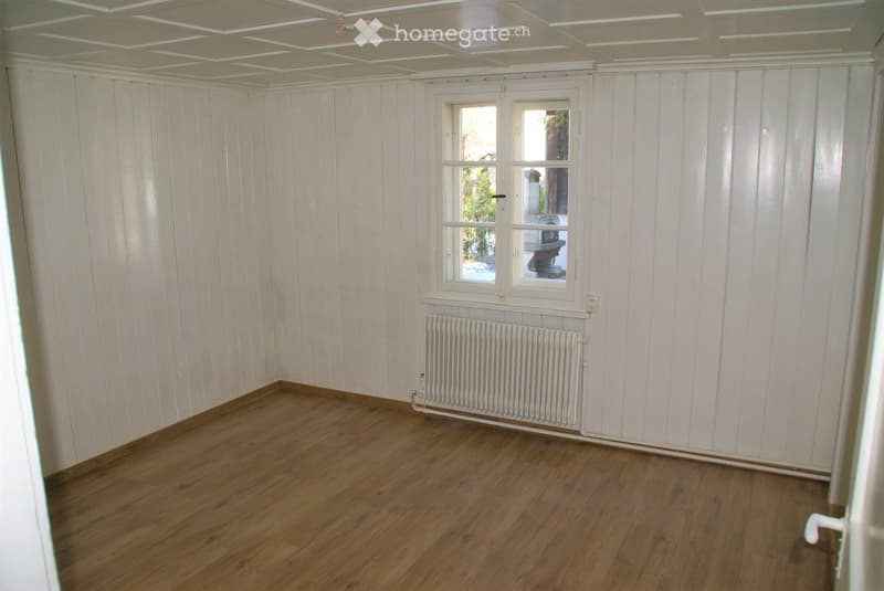 Empty room with wooden flooring, white paneled walls, a single window, and a radiator below it.