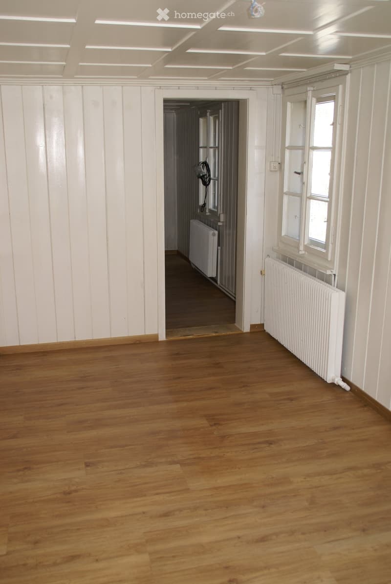 Empty room with wooden flooring, white paneled walls, and radiators visible. There is a doorway leading to another room with similar flooring and a radiator.