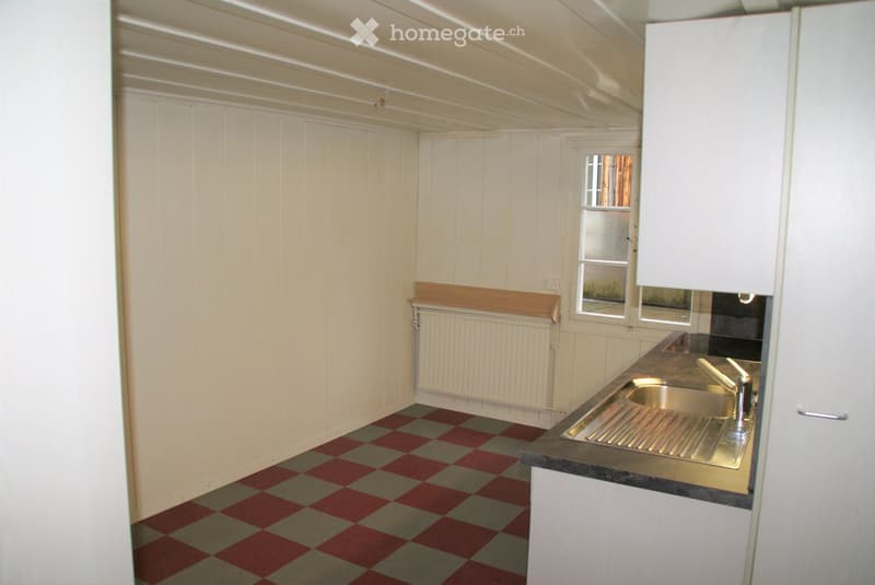 A small kitchen with white paneled walls, a checkered red and gray floor, a stainless steel sink, and white cabinets.
