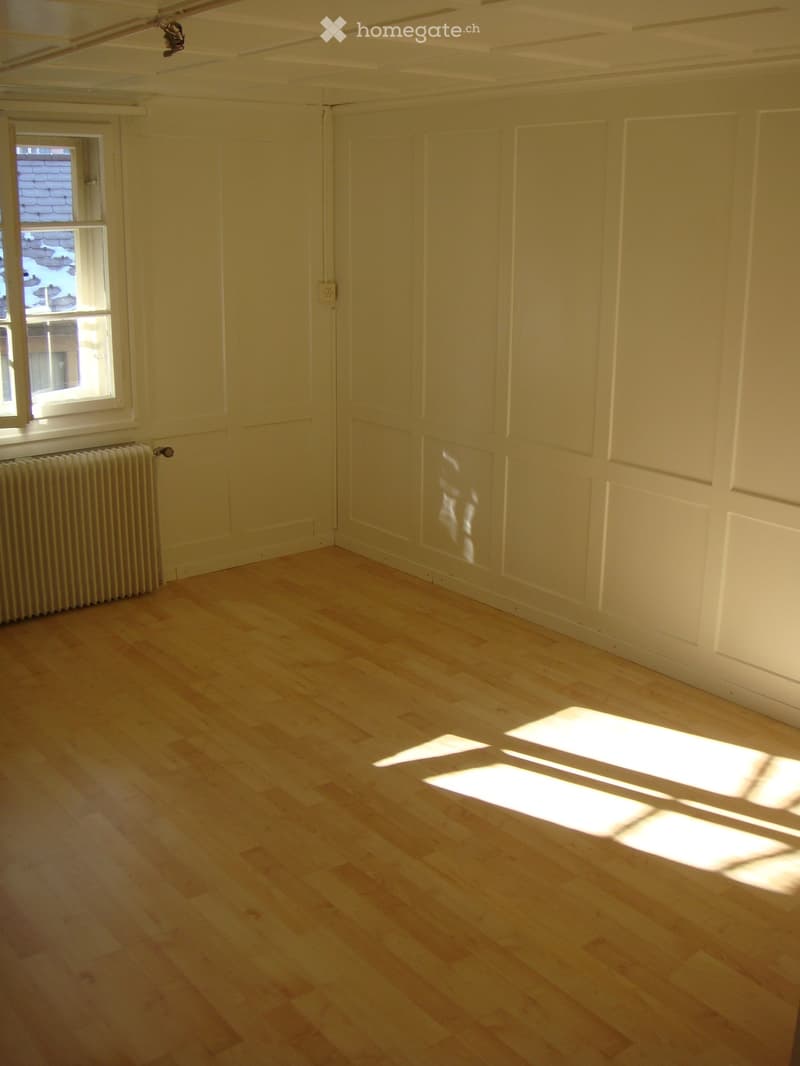 Empty room with light wooden flooring, white walls, and a single window letting in natural light. There is a radiator below the window and a closed door on the opposite wall.