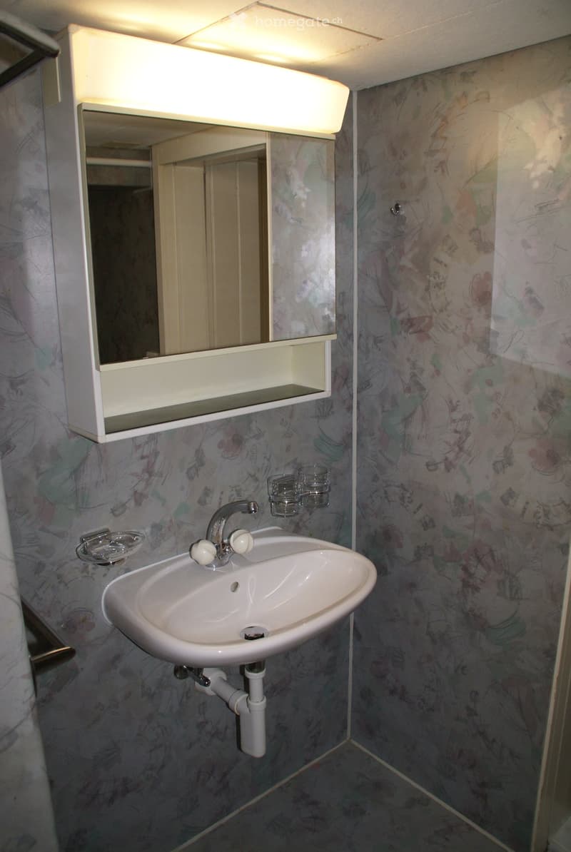 A small bathroom with a white sink, faucet, mirror, and two cups on a shelf above the sink. The walls are covered with floral patterned wallpaper.
