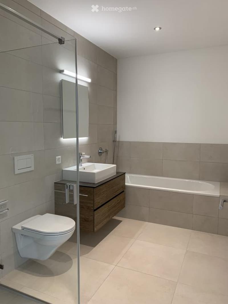 Modern bathroom with a white sink, toilet, bathtub, and a large mirror. The space features light-colored tiles and a sleek wooden cabinet under the sink.