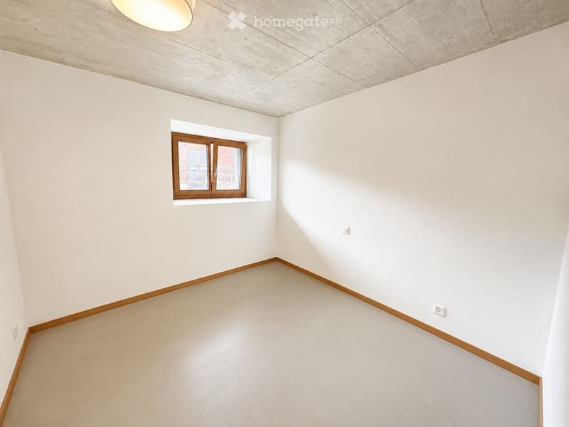 Empty room with light-colored walls, wooden baseboards, and a single window with wooden frame. The floor is a light beige color.