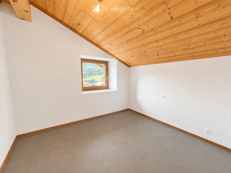 Empty room with light-colored walls, wooden trim, carpeted floor, and a single window with wooden frame showing a view of greenery outside.