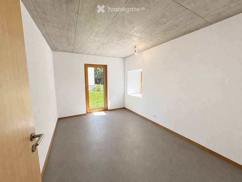 Empty room with white walls, grey carpeted floor, wooden trim, and a large wooden door leading outside. A small window with a white frame is visible on the right wall.
