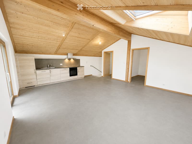 Open, unfurnished room with exposed wooden beams, white walls, grey carpet, and a small kitchen area with cabinets and appliances.