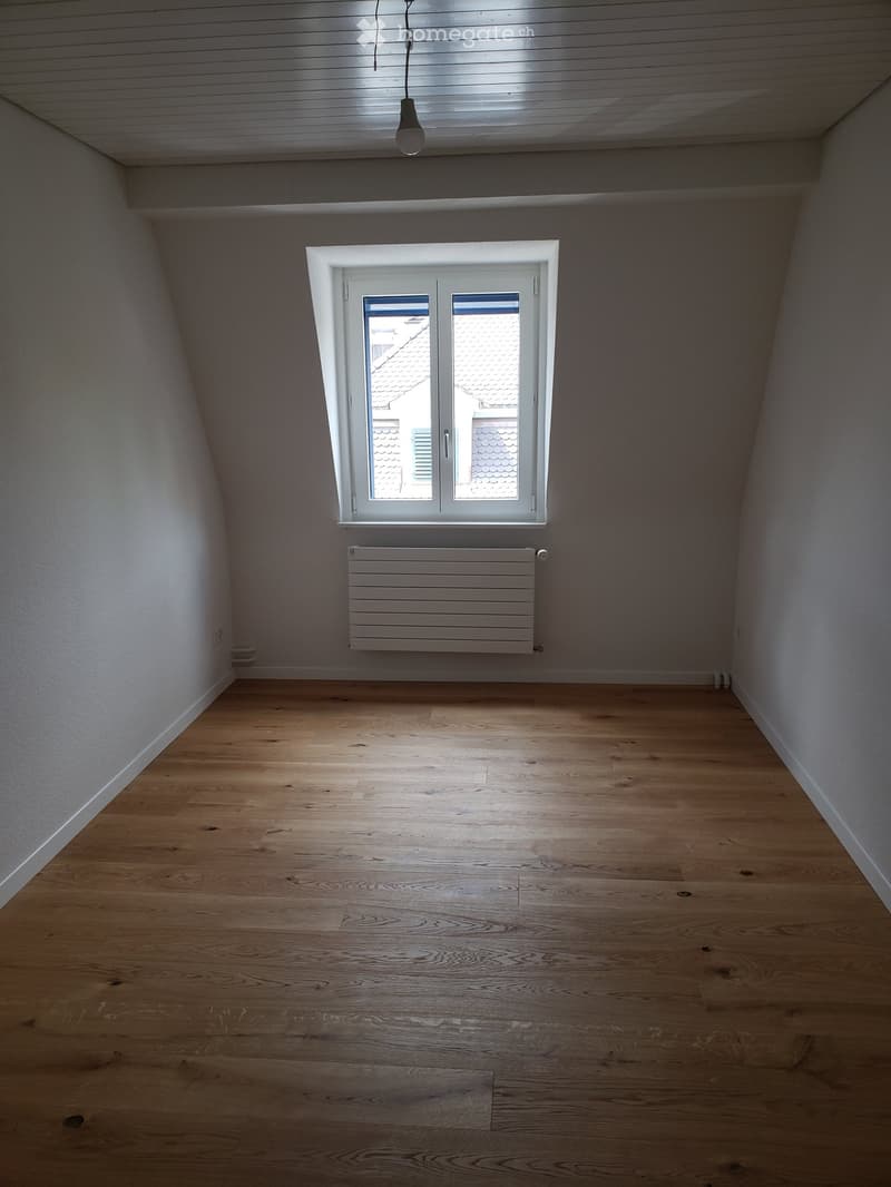 Empty room with wooden flooring, white walls, a window with double doors, and a radiator below the window.