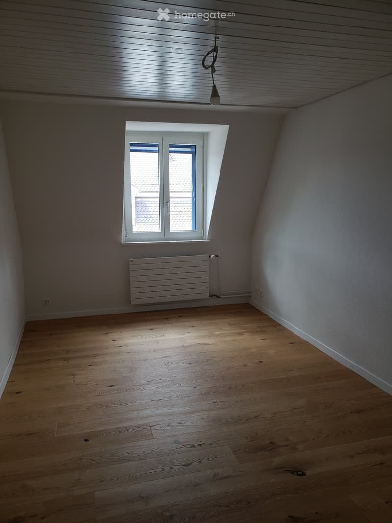 Empty attic room with wooden flooring, white walls, a single window, and a radiator. The ceiling has exposed wooden beams.