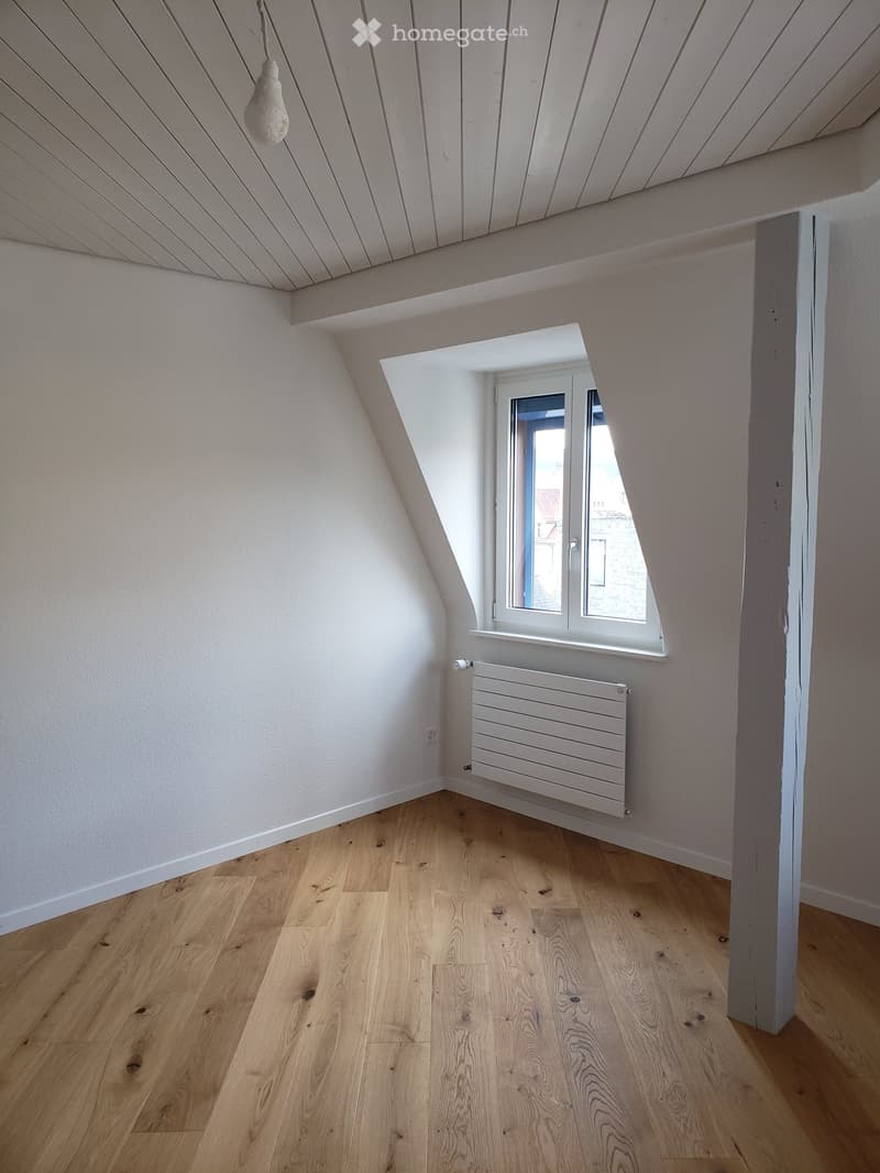 Empty attic room with wooden flooring, white walls, a window, and a radiator. The ceiling has exposed wooden beams.