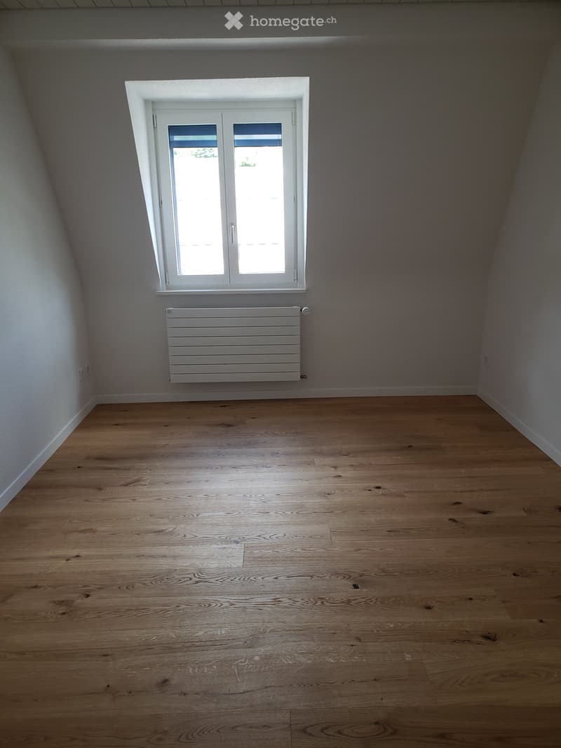 Empty room with wooden flooring, white walls, a window with white frame, and a white radiator below the window.