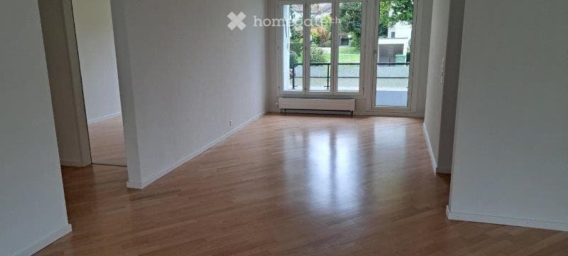 Empty room with wooden flooring, white walls, large window with view to garden, radiator below window.