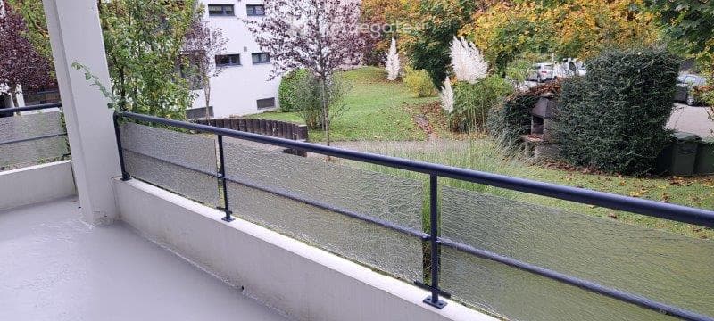 Balcony with glass railing, overlooking a green lawn and trees, with a building in the background.