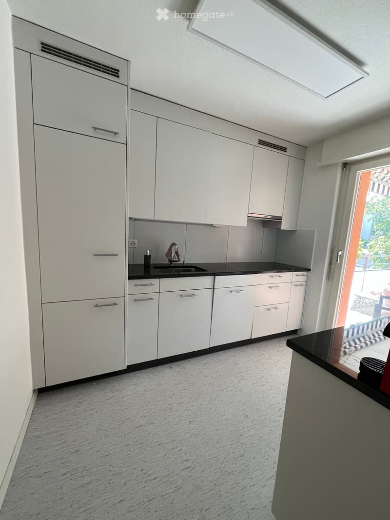 Modern kitchen with white cabinets, black countertop, and a sink. The floor is made of light-colored stone tiles.