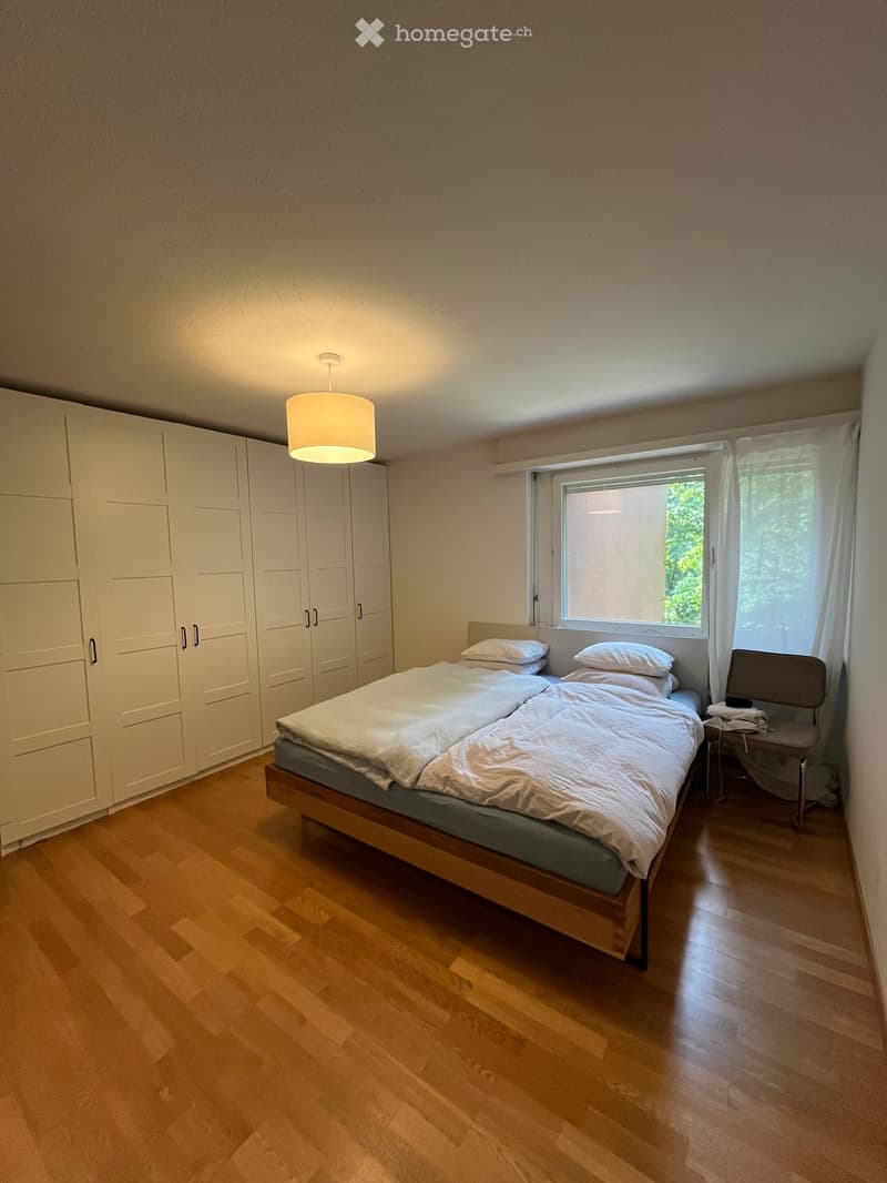 Bright bedroom with wooden flooring, a double bed with white bedding, a wardrobe, a chair, and a window with white curtains.