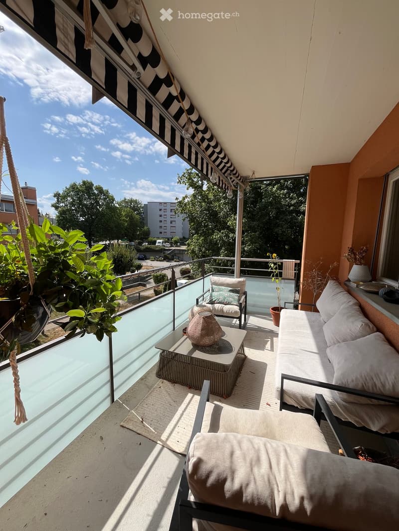 Balcony with a sofa, table, chair, vase, and potted plants, featuring a glass railing and a view of greenery and buildings.