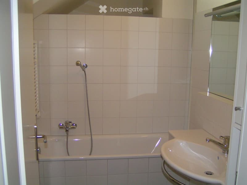 A small bathroom with a bathtub, a sink, and a mirror. The walls are tiled in light beige tiles, and the fixtures are modern and silver.