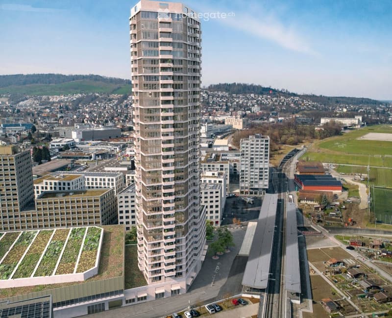 Aerial view of a modern urban area with a tall residential building, surrounded by other structures, green spaces, and a railway line.
